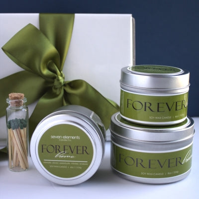 Three 'Forever' candles in metal tins with a bottle of matches and a gift box with a green ribbon on a white background.