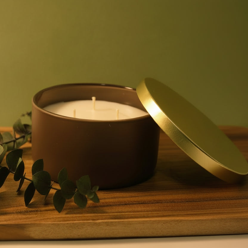 Candle in a brown tin with a gold lid on a wooden board against a green wall.